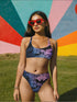 Model wearing a high-waisted pastel goth bikini set featuring a black bat and spiderweb print over an ombré pink and purple background. The two-piece swimsuit includes a scoop neck top and high-rise bottoms, styled outdoors in bright festival and poolside settings. Made of recycled fabric, soft, stretchy, and UV resistant. Available in XS to 3X. Aesthetic is gothic, spooky cute, and Y2K inspired.